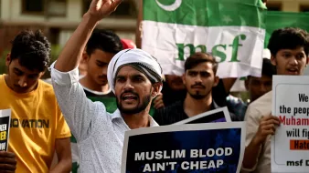 L'image montre un groupe de manifestants rassemblés, tenant des pancartes et des drapeaux. Au premier plan, un homme s'exprime avec passion, levant un doigt en l'air. Il porte une chemise claire et un turban. Les autres participants dans le fond semblent également engagés dans la manifestation, brandissant des affiches avec des messages. L'atmosphère semble intense et revendicative.