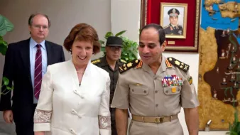 L'image montre une scène de rencontre entre plusieurs personnes dans un environnement officiel. Une femme souriante, vêtue d'une tenue claire, semble discuter avec un homme en uniforme militaire. Ce dernier porte des médailles et des insignias sur son uniforme. En arrière-plan, on peut voir un homme en tenue militaire additionnelle et une carte ou un tableau accroché au mur. L'ambiance semble amicale et détendue, suggérant un dialogue diplomatique ou militaire.
