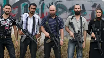 L'immagine mostra un gruppo di cinque persone, tutte in piedi e con armi da fuoco in mano. Quattro di loro hanno un aspetto maschile, mentre una è femminile. Sono vestiti in abiti casual e mostrano espressioni serie. Sullo sfondo si intravedono graffiti, creando un'atmosfera di tensione. L'ambiente sembra essere abbandonato o in degrado, con erba alta che circonda i soggetti.