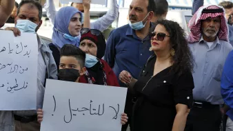 در این تصویر، گروهی از افراد در حال تظاهرات هستند. برخی از آنها ماسک به صورت دارند و تابلوهایی در دست دارند. یکی از تابلوها حاوی جمله‌ای است که به زبان عربی نوشته شده و به معنای "نه به تاخیر!" است. افراد در تصاویر شامل زنان و مردانی با پوشش‌های مختلف هستند، و برخی از آنها با چهره‌های جدی و مصمم به بیان خواسته‌های خود پرداخته‌اند. فضای کلی نشان‌دهنده‌ی اعتراض و تلاش برای بیان نظرات و مطالبات است.