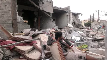 L'image montre une scène de destruction, probablement après un conflit ou une catastrophe naturelle. Au premier plan, un homme assis sur des matelas usés semble perdu dans ses pensées, entouré de débris. À l'arrière-plan, des bâtiments partiellement effondrés et des gravats jonchent le sol, témoignant des conséquences dévastatrices de l'événement. L'atmosphère est sombre et tragique, avec une sensation de désolation.