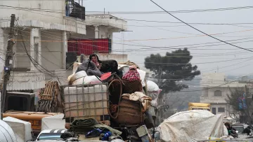 L'image montre une scène urbaine chargée, où des personnes sont perchées sur une pile de marchandises. Elles semblent se déplacer sur un véhicule surchargé, avec différents types de bagages et de matériaux. L'environnement est voilé par une lumière tamisée, peut-être due à un temps nuageux ou brumeux, et des câbles électriques traversent la scène. Les bâtiments en arrière-plan suggèrent un contexte urbain. Cette image évoque un sentiment de mouvement et de survie dans un espace urbain encombré.