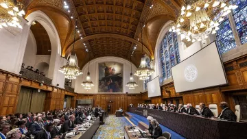 L'image montre l'intérieur d'une salle de tribunal, probablement une cour internationale. On peut y voir de nombreuses personnes assises, certaines en robes judiciaires. Au fond, des juges sont assis sur un banc élevé, entourés de décorations élégantes et de grandes fenêtres ornées de vitraux. La salle est richement décorée, avec des plafonds en bois et des lustres suspendus, créant une atmosphère solennelle et formelle. L'ensemble évoque un cadre judiciaire prestigieux.