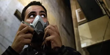 L'image montre un homme qui porte un masque à gaz. Son visage exprime une certaine vigilance, et il semble regarder autour de lui. L'environnement est urbain, avec des murs et des bâtiments en arrière-plan. L'éclairage donne une ambiance sombre et intense à la scène.