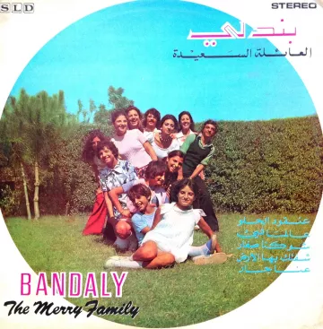 L'image représente une couverture de disque avec un groupe de personnes posant ensemble à l'extérieur, probablement dans un jardin. Ils semblent s'amuser et sourire, créant une ambiance joyeuse. Le nom du groupe, "BANDALY", et le titre "The Merry Family" sont présentés en bas, avec des inscriptions en arabe. En arrière-plan, on peut voir des arbres et une haie. L'ensemble évoque une atmosphère festive et conviviale.