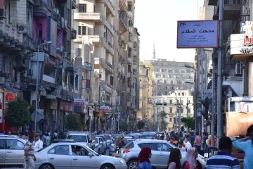 L'image montre une rue animée dans une ville, probablement une métropole. On peut voir de nombreux bâtiments de plusieurs étages qui bordent la route. Les voitures sont garées et circulent, créant un certain embouteillage. Une foule de personnes se déplace le long des trottoirs. Des panneaux de signalisation et des enseignes en arabe sont visibles, indiquant que cette scène se déroule dans un pays arabophone. L'atmosphère semble dynamique et vivante, typique d'une grande ville.