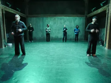 L'image montre un groupe de six personnes sur scène, dans un espace artistique éclairé d'une lumière verte. Elles semblent engagées dans une performance ou une répétition. Les membres du groupe portent des vêtements de différents styles, et se déplacent à divers endroits de la scène. L'arrière-plan est dépouillé, avec des murs en béton, ce qui crée une ambiance minimaliste et contemporaine.
