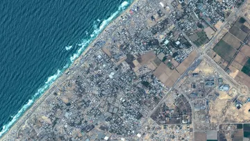 L'immagine mostra una vista aerea di un'area costiera. Si possono osservare le onde del mare che si infrangono sulla riva, evidenziando una linea di costa ben definita. L'area terrestre è caratterizzata da un mix di edifici e infrastrutture, con una disposizione varia di strade e terreni agricoli. I colori variano tra il blu dell'acqua, il verde delle aree coltivate e il grigio dei centri abitati e delle strutture urbane. L'immagine presenta un contrasto interessante tra le zone urbanizzate e quelle naturali.