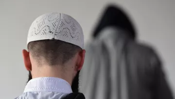 الصورة تظهر شخصًا يرتدي قبعة تقليدية بيضاء، مع ظهور جزء من رأسه وظهره. خلفه يوجد شخص آخر يرتدي غطاء رأس داكن مع معطف، مما يخلق تباينًا في الألوان. الجو العام للصورة يوحي بالهدوء والتركيز، وقد تكون في سياق ديني أو روحي.