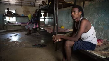 L'image montre un espace intérieur qui semble être une chambre ou un dortoir. On y voit plusieurs lits superposés et des personnes assises ou allongées. Un jeune homme est assis sur le bord d'un lit, regardant pensivement au loin. Il porte un débardeur et un short, et ses pieds sont nus. Les murs de la pièce sont nus et usés, avec des traces d'humidité, ce qui indique un environnement modeste et probablement surpeuplé. La lumière naturelle pénètre par une fenêtre, ajoutant une certaine ambiance à la scène.