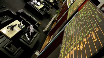 The image depicts a financial trading environment, likely within a stock exchange. In the foreground, there is a large electronic display board showing various financial data, such as stock prices and indices, with green and red text indicating changes in value. Below the board, there are several black leather seating areas where traders or visitors might be sitting. One individual, dressed in traditional attire, appears to be engaged with a computer or taking notes. The overall atmosphere suggests a busy and professional financial setting.