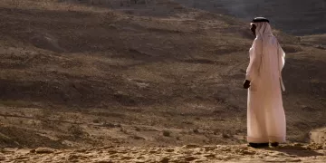 L'image montre une personne vêtue d'une tenue traditionnelle, probablement un kandora ou un dishdasha, se tenant debout sur un terrain désertique. En arrière-plan, on aperçoit un paysage aride avec des collines et des formations rocheuses. La lumière est douce, ce qui donne une ambiance calme et contemplative à la scène. La personne semble regarder au loin, peut-être en train de réfléchir ou d'admirer la vue panoramique du désert.