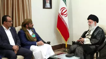 الصورة تُظهر ثلاثة رجال يجلسون في غرفة اجتماعات. في الخلفية، يوجد علم إيران وصورة كبيرة على الحائط. الرجل الموجود على اليمين يرتدي زيًا تقليديًا ويبدو أنه يتحدث مع الرجلين الآخرين، الذين يجلسون أمامه. يُظهر تعبيرهم اهتمامًا وتفاعلًا مع الحديث. الغرفة تبدو رسمية، مع أثاث بسيط وإضاءة دافئة.