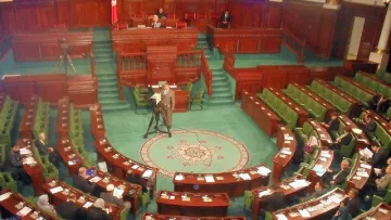 L'image montre l'intérieur d'un parlement, probablement un hémicycle de l'assemblée nationale. On peut voir plusieurs rangées de sièges verts disposés en arc de cercle, où des membres semblent assister à une session. Au centre, un tapis décoratif est visible, et à l'arrière-plan, on aperçoit une estrade pour les orateurs. Des caméras sont présentes, indiquant que la session est probablement diffusée. L'ensemble dégage une atmosphère formelle et politique.
