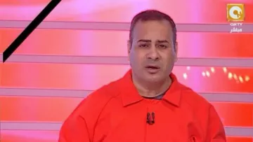 L'image montre un homme en train de parler devant un fond rouge avec des lignes lumineuses. Il porte une combinaison orange et semble être en train de faire une présentation ou de commenter quelque chose. Son expression faciale est sérieuse et il tient un stylo dans sa main. Le décor et son apparence suggèrent qu'il s'agit d'un animateur ou d'un commentateur à la télévision.
