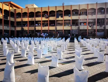 L'image représente un espace ouvert où de nombreux sacs blancs sont disposés en lignes. Ces sacs semblent être remplis de quelque chose, et le sol est constitué de pavés. En arrière-plan, on peut voir un bâtiment à plusieurs étages avec des balcons. Des vêtements sont suspendus à l'extérieur des balcons, suggérant une utilisation résidentielle de cet espace. La lumière du soleil éclaire la scène, créant un contraste entre les sacs blancs et l'environnement.