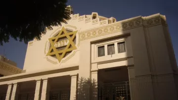 L'image montre un bâtiment architectural avec des éléments néo-classiques. La façade est ornée d'une étoile de David, symbolisant une influence juive. Les fenêtres sont encadrées de détails géométriques et il y a une entrée principale bien marquée. Le bâtiment est éclairé par la lumière du jour, ce qui met en valeur ses décorations. Des arbres sont visibles au premier plan, ajoutant une touche de verdure à la scène.