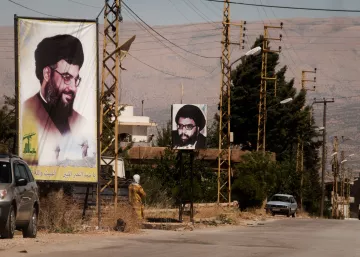 L'image montre une rue avec des panneaux d'affichage sur lesquels figurent des portraits d'un homme portant des lunettes et une barbe, associé à une ambiance rurale. On peut également voir des lignes électriques et quelques bâtiments en arrière-plan. À gauche, un panneau est en couleur et à droite, un autre est en noir et blanc. La scène semble être située dans une région montagneuse. C'est une illustration d'un espace public où des affiches politiques ou religieuses peuvent être observées.