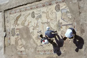 L'immagine mostra un antico mosaico, parzialmente scavato, con una varietà di animali e motivi decorativi. Si possono osservare figure di leoni, cervi e palme, che suggeriscono una rappresentazione di scene naturali. Due persone sono visibili nella parte inferiore, impegnate nel lavoro di pulizia o restauro del mosaico. L'illuminazione crea ombre sul pavimento, accentuando i dettagli del mosaico e l'atmosfera archeologica del sito.