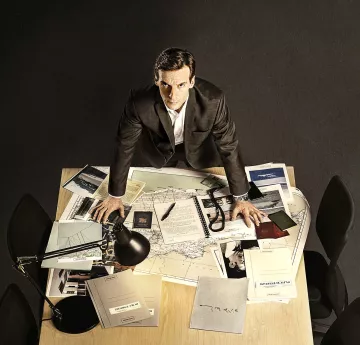 L'image montre un homme debout sur une table en bois, entouré de divers documents et d'objets. Il a une expression sérieuse et semble concentré. Sur la table, on peut voir des cartes, des notes, des dossiers et un lampadaire qui éclaire la scène. L'environnement autour de lui est plutôt sobre, avec des chaises noires et un sol sombre. L'ensemble donne une impression de travail acharné et de recherche.