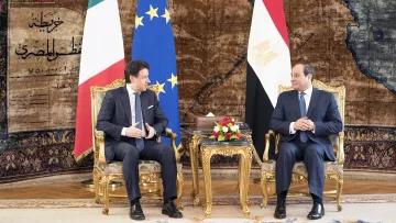 L'immagine mostra due uomini seduti su poltrone dorate in un ambiente elegante. Sullo sfondo si possono notare le bandiere dell'Italia e dell'Egitto, insieme a una bandiera dell'Unione Europea. Entrambi gli uomini stanno conversando e sembrano impegnati in una discussione formale, circondati da elementi decorativi e un tappeto elaborato. Sulle loro ginocchia ci sono bouquet floreali. L'atmosfera è di cordialità e ufficialità.