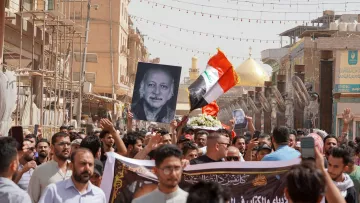 L'immagine mostra una folla di persone durante una manifestazione. Molti manifestanti tengono in alto una grande fotografia di un uomo, insieme a una bandiera irachena. Sullo sfondo si possono vedere elementi architettonici e due cupole dorate, che suggeriscono la presenza di un luogo sacro o importante. L'atmosfera sembra intensa e significativa, con i partecipanti che mostrano il loro sostegno o commemorazione attraverso slogan e simboli.