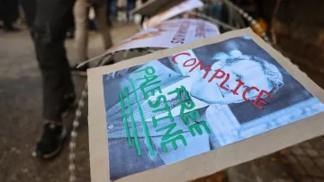 L'immagine mostra un cartello posizionato su una recinzione, con il volto di una persona ritratta in bianco e nero. Sotto l'immagine ci sono delle parole scritte con pennarelli colorati: "COMPLICE" e "FREE PALESTINE". Il contesto sembra essere un evento di protesta, con altri cartelli visibili in background e una folla di persone. L'atmosfera è quella di un'espressione di dissenso e di richiesta di libertà per la Palestina.