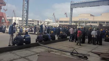 L'image montre un groupe de nombreux travailleurs rassemblés dans un espace extérieur, probablement dans un port ou un chantier naval. Les personnes sont vêtues de tenues de travail et portent des casques de sécurité jaunes. Certains d'entre eux sont assis sur le sol, tandis que d'autres sont debout, discutant ou attendant. L'environnement semble industriel, avec des structures métalliques et des équipements visibles en arrière-plan. Le ciel est partiellement nuageux.