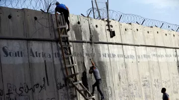 La imagen muestra a dos personas subiendo una pared de concreto alta. Una de ellas está en la parte superior, mientras que la otra está en una escalera de madera, intentando escalar. En la pared hay alambres de púas en la parte superior y también se pueden ver algunas inscripciones y grafitis en varios idiomas. El ambiente parece ser tenso y el contexto sugiere una situación de conflicto o segregación.