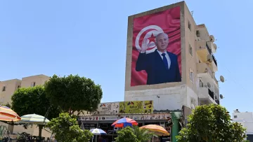 L'image montre un bâtiment avec une grande fresque représentant un homme, probablement un dirigeant, qui fait un geste de salutation. En arrière-plan, on peut voir un drapeau tunisien. Au premier plan, il y a des parasols colorés et des arbres, ainsi qu'une scène de vie qui pourrait inclure des personnes assises ou se déplaçant. Le ciel est clair et ensoleillé, ce qui donne une atmosphère lumineuse à la scène.