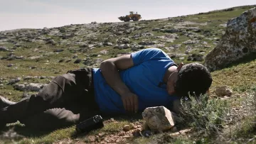 Nell'immagine si vede un uomo sdraiato su un prato verde, con il viso rivolto verso il terreno. Indossa una maglietta blu e pantaloni scuri. Accanto a lui ci sono alcune rocce. Sullo sfondo, si scorge un bulldozer che lavora su un terreno collinare. La scena trasmette un senso di tranquillità, ma anche di inquietudine a causa della presenza del macchinario che sottolinea il contrasto tra l'uomo e l'ambiente circostante.