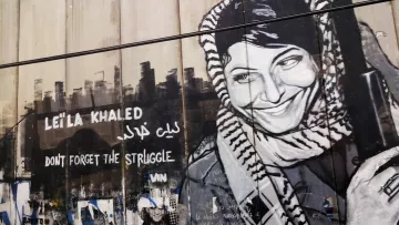 L'image présente une fresque murale représentant Leïla Khaled, une figure emblématique de la résistance palestinienne. Elle est dépeinte avec un sourire, portant une écharpe typique et tenant un fusil. À côté de son portrait, on peut lire des inscriptions en anglais et en arabe, telles que "Don't forget the struggle" et son nom. L'arrière-plan est constitué d'un mur, accentuant le message de lutte et de mémoire associé à son image. Des graffitis colorés et des marques diverses décorent également la surface, ajoutant à l'atmosphère militante et artistique du lieu.