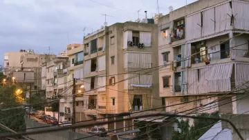 L'image montre un ensemble d'immeubles résidentiels dans une zone urbaine. Les bâtiments semblent anciens et sont de plusieurs étages, avec des balcons où l'on peut voir des vêtements étendus. La lumière du soir crée une atmosphère douce, et des fils électriques traversent l'image. On aperçoit également des voitures garées le long de la rue en contrebas. L'ensemble donne une impression de vie citadine typique, avec un mélange de modernité et de caractère ancien.