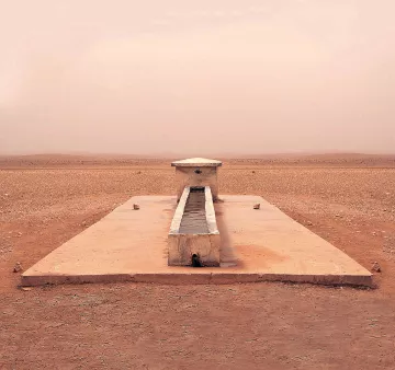 L'image montre une structure isolée située dans un paysage désertique. Au centre, il y a un long bassin en pierre, entouré par une surface plane et aride. Le sol est de couleur beige et semble sec et craquelé, tandis que l'atmosphère est légèrement brumeuse, ce qui donne une impression de chaleur et d'immensité désertique. Au loin, on aperçoit des collines ou des dunes qui se fondent avec le ciel dans une teinte douce et uniforme. L'endroit dégage une sensation de tranquillité et de solitude.