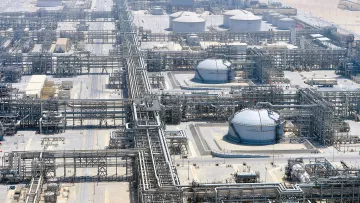 La imagen muestra una vista aérea de una planta industrial, probablemente relacionada con la producción o el refinado de petróleo. Se pueden observar grandes tanques de almacenamiento, tuberías extensas que se entrelazan por todo el área y estructuras de soporte. El entorno es desértico, con un paisaje árido al fondo, lo que sugiere que esta instalación podría estar ubicada en una región donde la extracción de recursos naturales es común. La organización y complejidad de las tuberías y tanques reflejan la sofisticación de la infraestructura industrial.