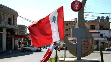 L'image représente un paysage urbain ensoleillé au Liban. On y voit un drapeau libanais flottant au vent, avec une grande emblème en métal en arrière-plan. Cet emblème est orné de symboles et de signification culturelle, illustrant probablement l'identité locale ou nationale. Les bâtiments autour semblent typiques de la région, avec des lignes électriques visibles et un ciel clair en arrière-plan.