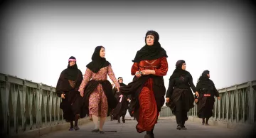 L'image montre un groupe de femmes marchant sur un pont. Elles portent des vêtements traditionnels, principalement des robes longues et des foulards. La scène semble être en extérieur, avec un ciel clair en arrière-plan. Les femmes ont des expressions déterminées et se déplacent ensemble, ce qui dégage une impression d'unité et de force.