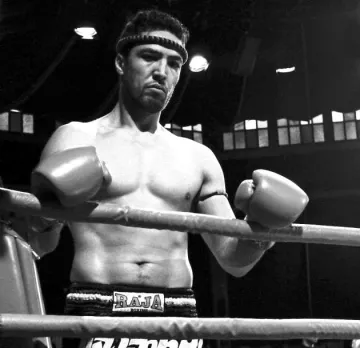 L'image montre un boxeur debout dans un ring. Il est torse nu et porte un bandeau autour de la tête. Ses poings sont protégés par des gants de boxe. Son expression semble concentrée et déterminée. Le style de l'image est en noir et blanc, ce qui lui donne une ambiance nostalgique et intemporelle. Le ring et l'arrière-plan suggèrent un environnement de compétition.