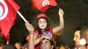 L'image montre une scène de célébration en Tunisie. Une jeune fille, vêtue d'une robe colorée, est assise sur les épaules d'un homme qui sourit. Ils brandissent des drapeaux tunisiens. En arrière-plan, on aperçoit une foule joyeuse, créant une atmosphère festive. Les expressions des visages reflètent la joie et l'engagement.
