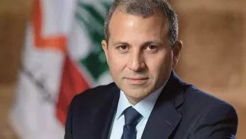 الصورة تظهر رجلًا يرتدي بدلة رسمية داكنة، مع قميص أبيض وروابط عنق زرقاء. يجلس بشكل مشي هادئ وملامحه تظهر الثقة. في الخلفية، تتواجد علم لبنان، مما يشير إلى احتمال ارتباطه بالسياسة أو الحكومة في لبنان. الجو العام للصورة يعكس الرسمية والجدية.