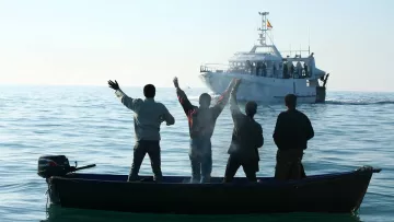 The image depicts four individuals in a small boat on calm waters, facing away from the camera. They are raising their arms, seemingly in a gesture of celebration or greeting. In the background, a larger boat is approaching, with several people visible on it. The scene is set against a clear sky, suggesting a peaceful day on the water.