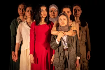 L'image montre un groupe de sept femmes, chacune portant des vêtements différents. Elles se tiennent en rang, avec des expressions sérieuses sur leurs visages. La femme au centre, vêtue d'une robe rouge, attire particulièrement l'attention. Les autres femmes, portant des vêtements variés allant de styles modernes à plus traditionnels, affichent une diversité de couleurs et de textures. L'éclairage met en valeur leurs visages et leurs postures, créant une atmosphère à la fois forte et émotive.