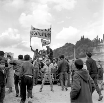 L'image montre une scène de rue animée, probablement lors d'une manifestation ou d'un rassemblement. Des groupes de personnes, en majorité des jeunes, se rassemblent autour d'une voiture. Certains tiennent des drapeaux ou des banderoles sur lesquelles des messages sont affichés, exprimant des revendications. L'atmosphère semble dynamique et chargée d'émotion, avec des visages montrant diverses réactions. En arrière-plan, on aperçoit un paysage urbain avec des arbres et des bâtiments. Le noir et blanc de la photo accentue le caractère historique et sérieux de la scène.