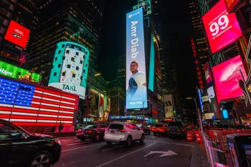 The image depicts a vibrant scene in Times Square, New York City, at night. The area is illuminated by numerous bright advertisements and digital billboards. Prominently featured is a large advertisement for Amr Diab, an acclaimed musician, which invites viewers to listen to his music. There is an American flag displayed on one of the buildings, and several vehicles are seen navigating the busy street. The atmosphere is lively, characteristic of this iconic urban location.