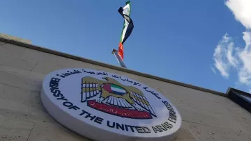 L'image montre un bâtiment avec un panneau qui indique qu'il s'agit de l'ambassade des Émirats arabes unis. Au-dessus du panneau, on peut voir le drapeau des Émirats arabes unis flottant. Le ciel est clair et légèrement nuageux, ce qui donne une belle arrière-plan à la scène.