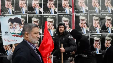 L'image montre une manifestation avec des affiches en arrière-plan affichant des personnages aux visages provocateurs, probablement des figures politiques, portant des coiffures spécifiques. Des gens tiennent des pancartes et des drapeaux, tandis qu'un homme sur le devant semble exprimer un message clair de désaccord ou de protestation. L'ensemble de la scène dégage une ambiance de contestation, soulignant des tensions politiques.