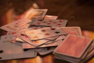 L'image montre un tas de cartes à jouer éparpillées sur une surface. On peut voir différents types de cartes, y compris des cartes de cœur, de carreau, de trèfle et de pique. Certaines cartes semblent retournées, tandis que d'autres sont face visible, montrant leurs motifs et valeurs. L'éclairage apporte une ambiance chaleureuse et met en valeur la texture des cartes.