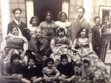 L'image présente un groupe de personnes, probablement une famille ou une communauté, posant ensemble dans un décor intérieur. On peut voir des hommes et des femmes de différentes générations, ainsi que des enfants, vêtus de tenues traditionnelles. L'atmosphère semble conviviale et chaleureuse. L'arrière-plan est orné de motifs, ajoutant une touche historique à la scène.
