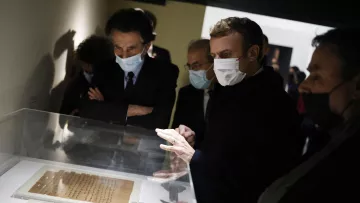 L'image montre un groupe de personnes, dont une figure centrale qui semble être un homme en train d'observer des documents exposés dans une vitrine. Ils portent tous des masques, probablement en raison de mesures sanitaires. L'environnement est sombre, avec des murs clairs, et il y a un intérêt évident pour les objets présentés. Les autres personnes dans le cadre semblent également attentives et engagées dans la discussion.