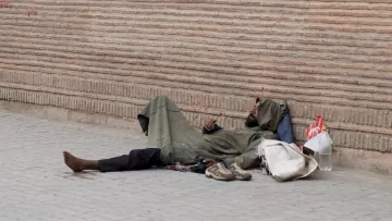 الصورة تظهر شخصًا ممددًا على الأرض، يرتدي ثيابًا بسيطة وفضفاضة. يبدو أنه في حالة استرخاء أو نوم. بجانبه بعض الأغراض مثل حذاء وحقيبة. الخلفية تتكون من جدار مصنوع من الطوب، مما يضيف إلى الأجواء الحضرية للصورة. المشهد يعكس حالة التحدي والمعاناة في الحياة اليومية لبعض الأفراد.